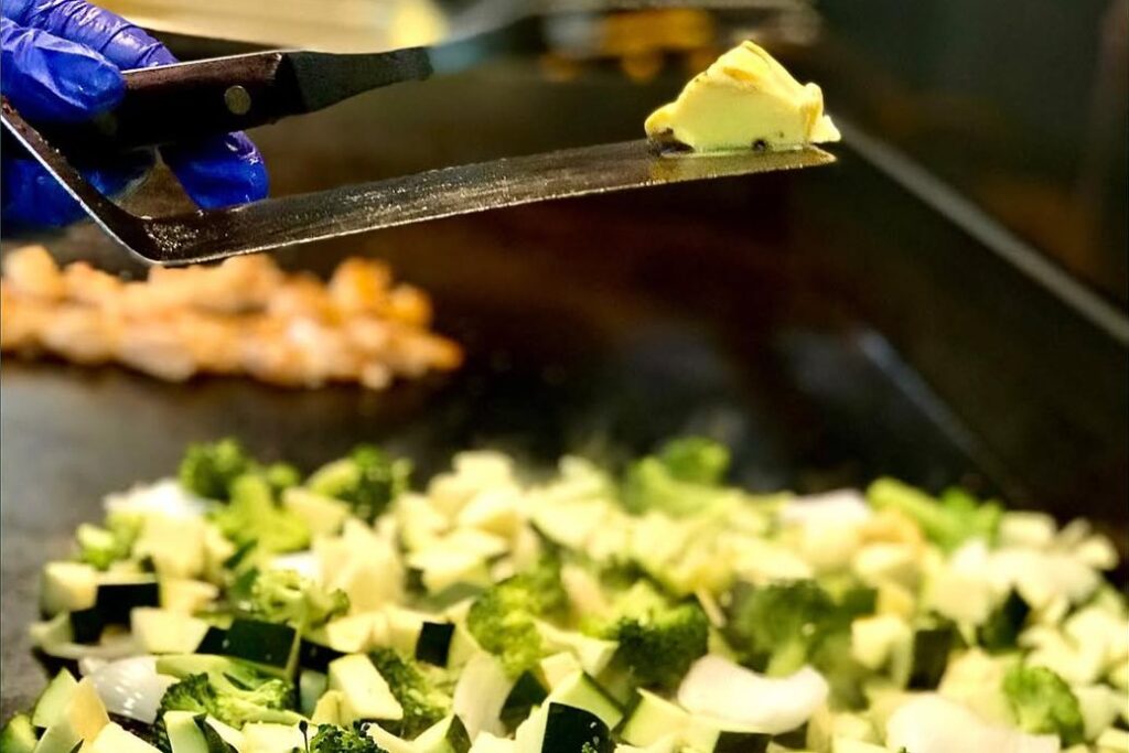A Bamboo Hibachi cook adding butter to diced zucchini, onion and broccoli on the flat-top grill, with shrimp cooking behind