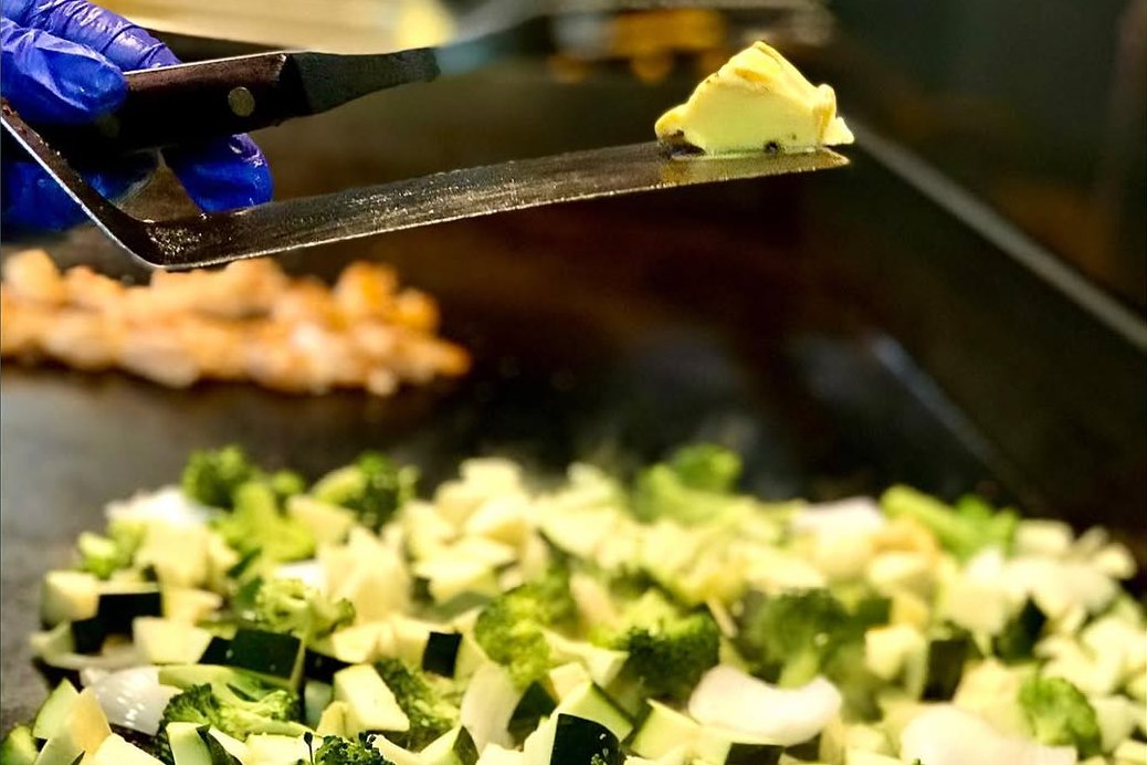 A Bamboo Hibachi cook adding butter to diced zucchini, onion and broccoli on the flat-top grill, with shrimp cooking behind